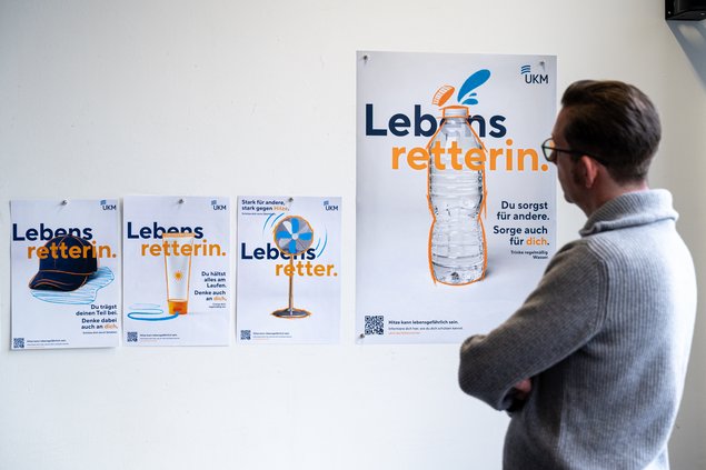 An einer Wand hängen vier Plakate. Das rechte ist größer als die anderen. Ein Mann steht mit verschränkten Armen davor und betrachtet diese.